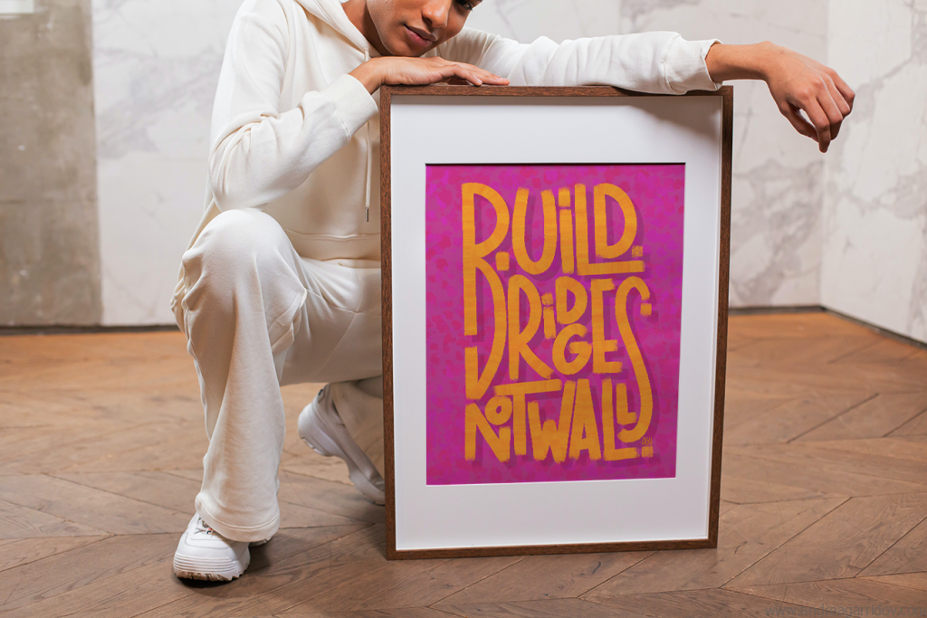 Persona apoyada en un marco de poster que dice Build bridges not walls en letras mezcladas naranja, con fondo magenta con textura en otros tonos.